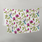 Magnolia Dreams throw blanket hanging on a line, featuring pink magnolia blossoms, colorful hummingbirds, butterflies and green leaves on a white background
