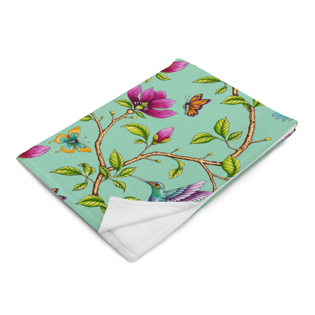 Magnolia Dreams throw blanket in Vista Blue, folded to showcase pink magnolia blossoms, colorful hummingbirds, butterflies and green leaves