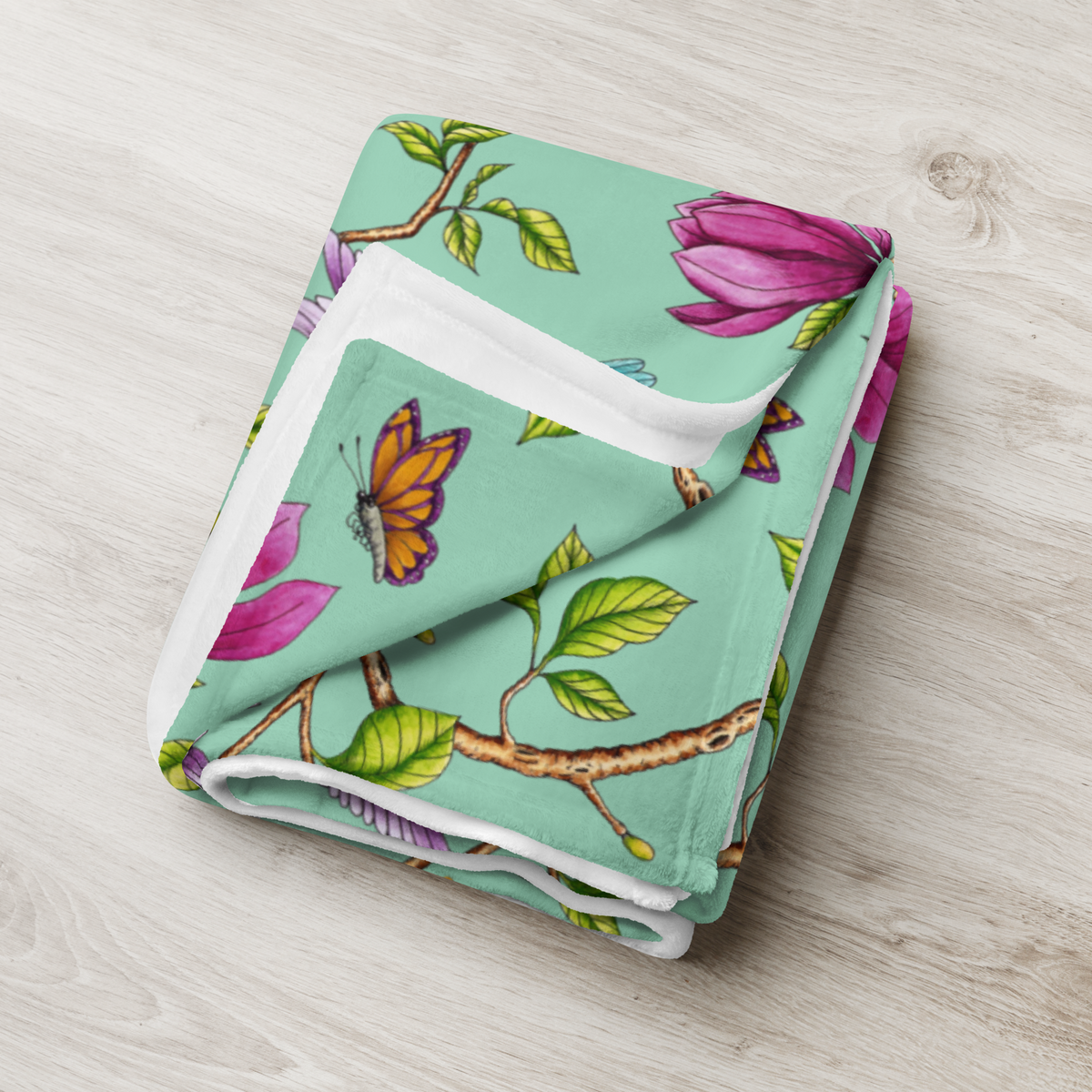 Folded Magnolia Dreams throw blanket in Vista Blue, featuring pink magnolia blossoms, colorful butterflies and green leaves