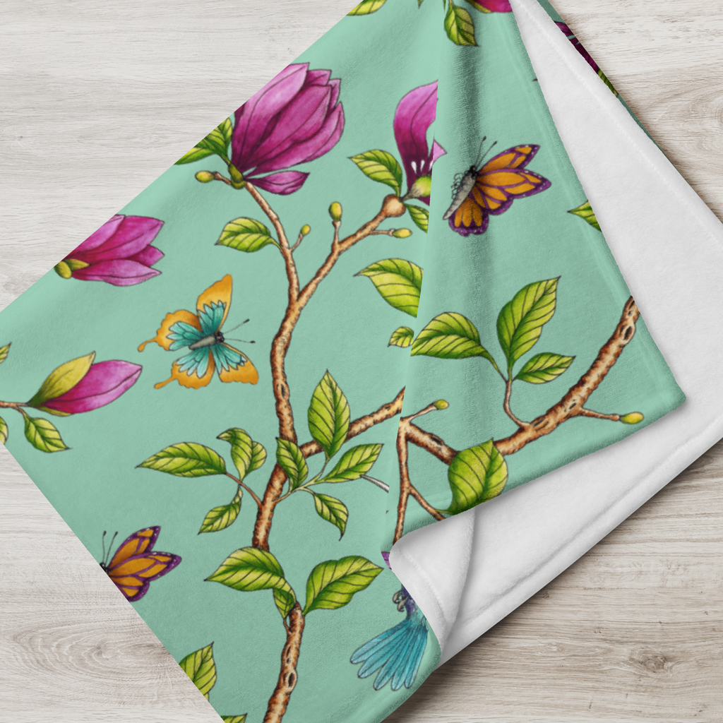 Close-up of a Magnolia Dreams throw blanket in Vista Blue, featuring pink magnolia blossoms, colorful butterflies and green leaves