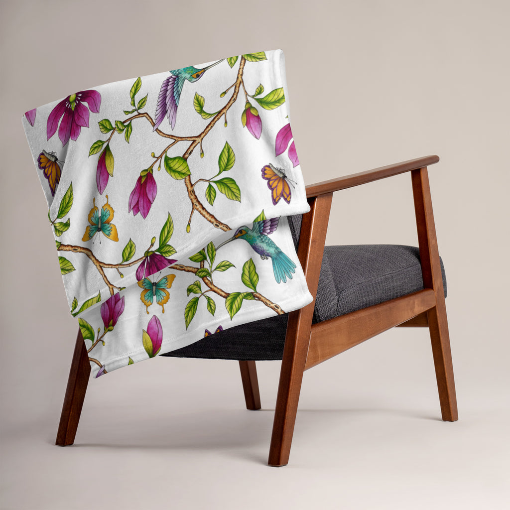 Magnolia Dreams throw blanket draped over a chair, featuring pink magnolia blossoms, colorful hummingbirds, butterflies and green leaves on a white background