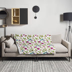 Garden of Eden throw blanket draped over a sofa, featuring pink magnolias, colorful hummingbirds and butterflies on a white background
