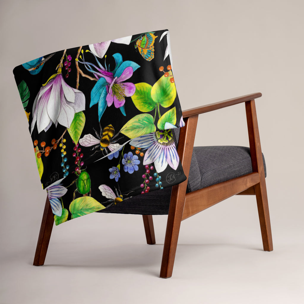 Garden of Eden throw blanket draped over a chair, featuring colorful exotic flowers, a kingfisher, bumblebees and green leaves on a black background