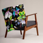 Garden of Eden throw blanket draped over a chair, featuring colorful exotic flowers, a kingfisher, bumblebees and green leaves on a black background