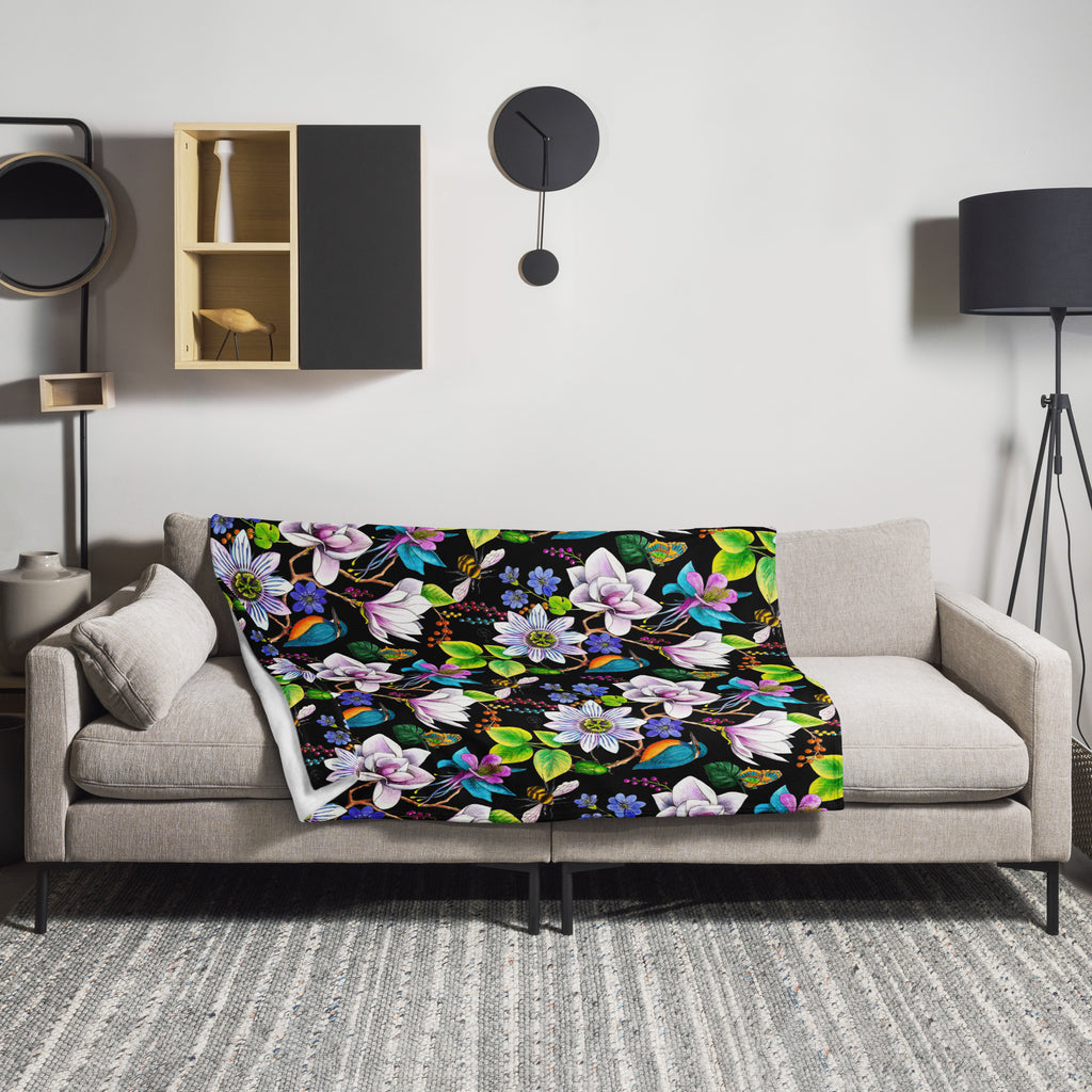 Garden of Eden throw blanket draped over a sofa, featuring colorful exotic flowers, kingfishers, butterflies and lush green leaves on a black background