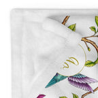 Close-up of a Magnolia Dreams throw blanket featuring a colorful hummingbird, pink magnolia blossoms and green leaves on a white background